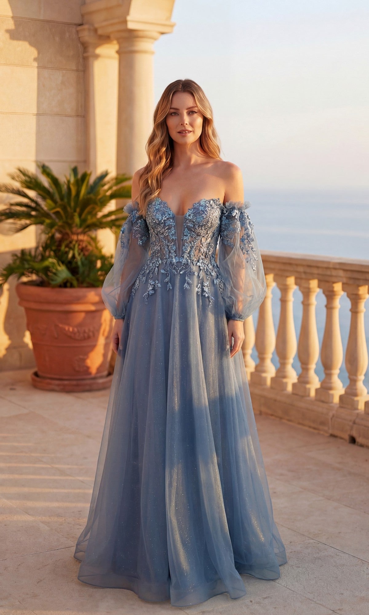 A woman in the Chicas C358 long formal dress in slate blue, featuring floral embroidery and a corset bodice, stands on a terrace overlooking the sea at sunset.